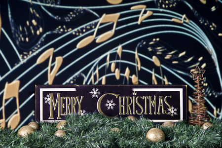 A black and gold Merry Christmas sign on a bed of garland and gold bulbs against a backdrop of musical staff and notes.の写真素材