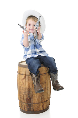 An adorable 2-year-old cowboy happily sitting on an old barrel with a toy gun in each hand.  On a white background.の写真素材
