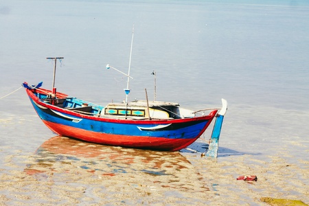 A small red blue Vietnamese fishing boat is lying on the beach. The beach is soon covered by the...