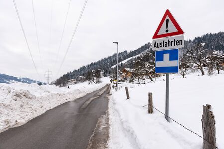 Red triangle road traffic warning sign - Other Danger with text Ausfahrten which means Exits. Snow covered road, trees and mountains. Austriaの写真素材
