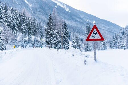 Black ice danger - traffic sign slippery road warning, winter services, Schladming Dachstein. Snow covered road, trees and mountains. Austria, Europeの写真素材