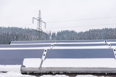 Photovoltaic panels partly covered with snow, installed on the roof of the house. In the background an element of the power grid - a high voltage power pole with overhead wires.の写真素材