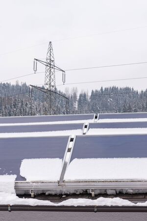 Photovoltaic panels partly covered with snow, installed on the roof of the house. In the background an element of the power grid - a high voltage power pole with overhead wires.の写真素材