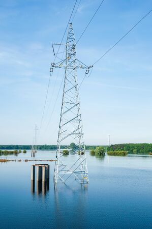 The high voltage power lines on water. Blue sky and green trees during river flood via pylon tower. Sunny spring day.の写真素材