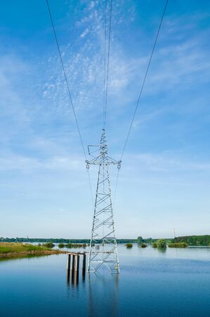 The high voltage power lines on water. Blue sky and green trees during river flood via pylon tower.の写真素材