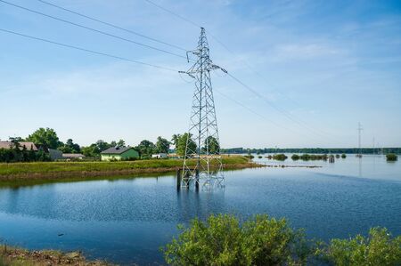 The high voltage power lines on water. Blue sky and green trees during river flood via pylon tower. Sunny spring day.の写真素材