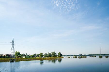 The high voltage power lines on water. Blue sky and green trees during river flood via pylon tower.の写真素材