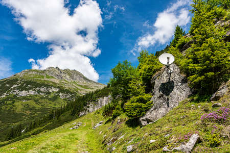 Satellite antenna mounted on a rock, around the green mountains. Beautiful summer mountain landscape - blue sky with white clouds.の写真素材