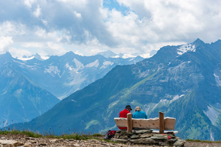 Two older people, tourists, rest on a bench in the mountains. Around them there is a view of the majestic snow-covered Alps.の写真素材