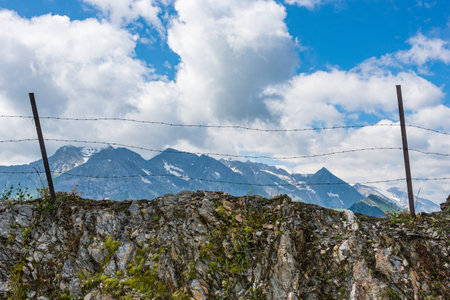 Barbed wire separating the mountain landscape. In the foreground there are solid stones and mountain plants, flowers. In the background high mountains, white clouds and blue sky.の写真素材