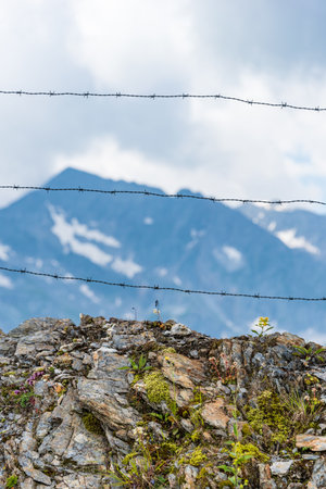 Barbed wire separating the mountain landscape. In the foreground there are solid stones and mountain plants, flowers. In the background high mountains, white clouds and blue sky.の写真素材