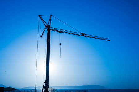 Silhouette, outline of the crane on the construction site against the blue sky and the sea. A fragment of the building, ribbed walls and reinforcement visible in the foregroundの写真素材