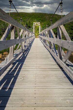 Long wooden plank suspension hiking bridge with cloudy skies and green forested hills in the...