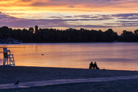 Couple sitting by the water with orange and violet morning sky reflection, sunrise on Dnipro river, Kyiv, Ukraineの写真素材