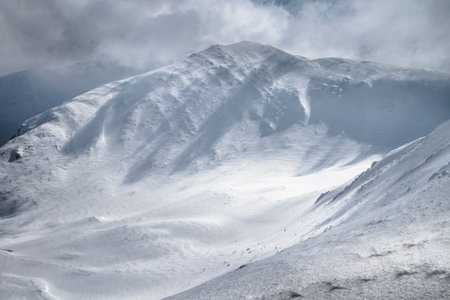 A large white rocky mountain with a dark couloir is covered with snow among the foggy clouds before...