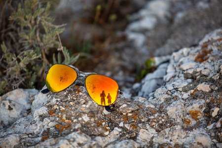 A pair of sunglasses bathed in orange light on a sea stone, with the reflection of tourists in them