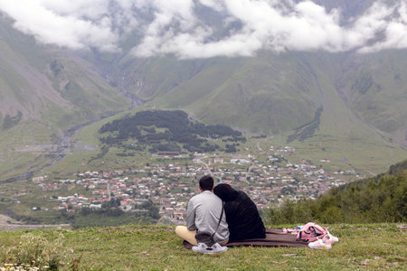 Serenity in the Caucasus: Two figures gaze at misty peaks and a town sprawled beneath rolling hills....