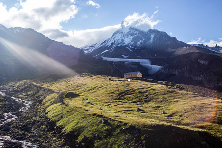 Kazbek's majesty: Sunbeams illuminate a lone shelter on verdant slopes, glacier-clad peak pierces...