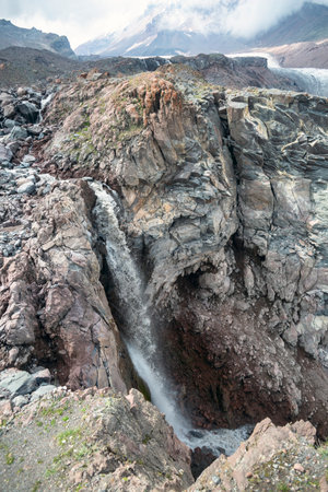 A cascading waterfall plunges down a rocky cliff, surrounded by a dramatic mountain landscape. A...
