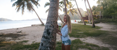 Beautiful young woman in a blue T-shirt and shorts standing near a palm tree on the beach.