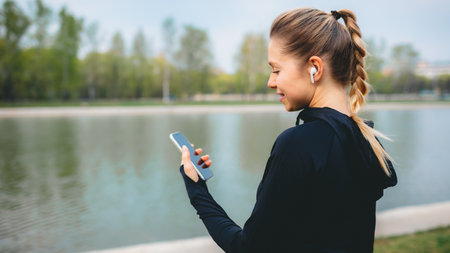 Young smiling girl making sport and running in the park using her phone to listen to the music with...