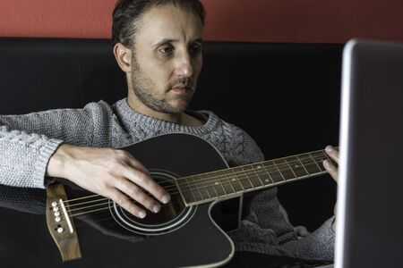 Man playing acoustic guitar in his room lying on the bed with the computerの写真素材