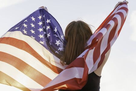 Beautiful young woman backwards during sunset holding the United States flag in the field celebrating United States Independence Day. July 4thの写真素材
