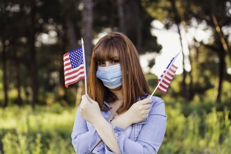 girl in protective medical mask with flags of United States of America in hands in nature. July 4 United States Independence Day. Health protection, safety and pandemic conceptの写真素材