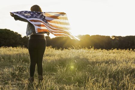 Beautiful young woman during sunset holding the United States flag in the field celebrating United...