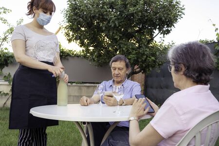 A waitress in a medical mask serves drinks to the tables to seniors outside the coronavirus outbreak lifestyle. selective focusの写真素材