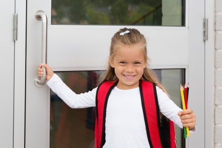 A Little Preteen Schilgirl Holding a Handle of a School Door Showing her Pencilsの写真素材
