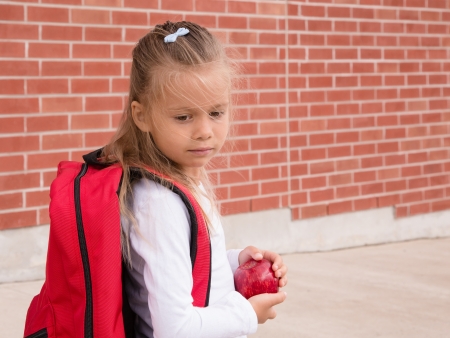 Young Schoolgirl with red backpack standing against brick school wall, holding an appleの写真素材