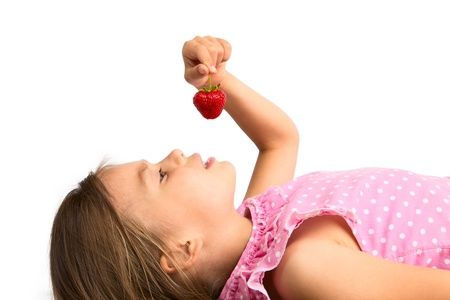 Beautiful younge girl in polka dot dress lying while holding a strawberry above her mouth, isolatedの写真素材