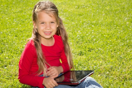 A cute schoolgirl sitting on a green lawn in summer park, holding  tablet computerの写真素材