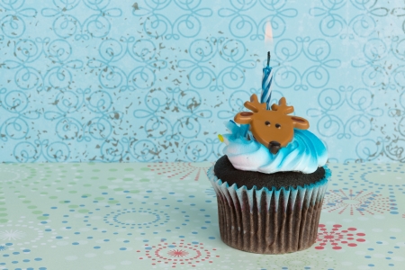 A colorful cupcake with toy deer on top and burning holiday candle inserted, against winter-themed backgroundの写真素材