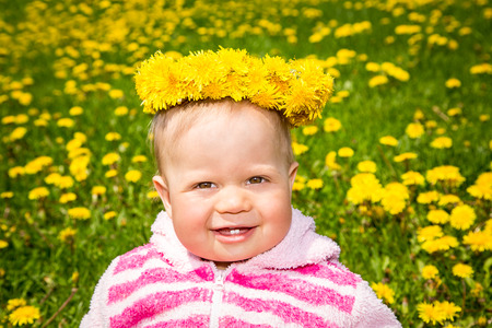 Cute baby girl  in a dandelion field, touching her wreath of dandelions and smiling happilyの写真素材
