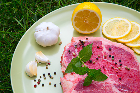 A cut of raw t-bone steak meat, with garlic, lemon and black peppers on a dish, on a fresh grassの写真素材