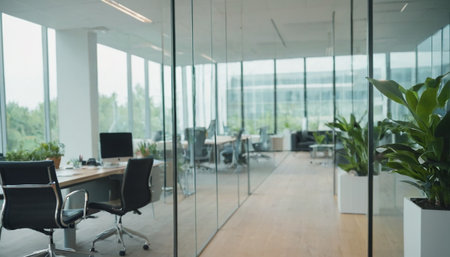 A and modern office interior characterized by sleek glass partitions that allow light to flow freely. Large windows provide ample natural illumination and a view of the surrounding greenery. Minimalist desks with computers and ergonomic chairs are arranged in a layout. Vibrant green potted plants add a refreshing natural to the clean and bright workspace, fostering a calm and productive environment.の素材