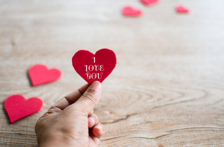 Concept Valentine's day, man's hand holding a red heart symbol, letters I love you white, and red heart symbol blur placed on a wooden table. Close-up, selective focus, copy space for design, and textの写真素材