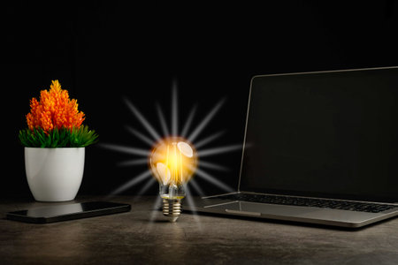A bright light bulb on work table. Concept of a startup that drives the economy, finance, banking. Wireless headphones in the working room. Laptop are black blank screen. Blurred, black backgroundの写真素材