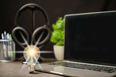 A bright light bulb and laptop are black blank screen on work table. Concept of a startup that drives the economy, finance, banking. Wireless headphones in the working room. Blurred, black backgroundの写真素材
