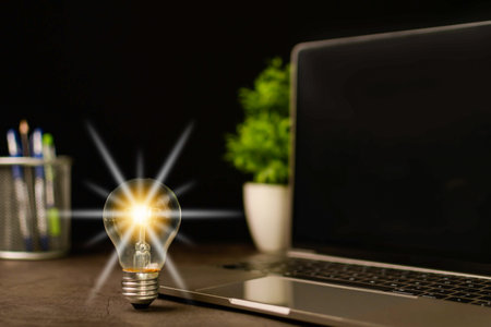 A bright light bulb and laptop are black blank screen on a work table. Concept of a startup that drives the economy, finance, banking. Bright lights give hope to businessmen. Blurred, black backgroundの写真素材