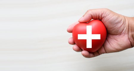 A red cross is a symbol of medical health. Hand holding a heart and on a wooden desk. Copy space for the text and contents. red cross CPR, World heart day, world health day, blood organ donationの写真素材