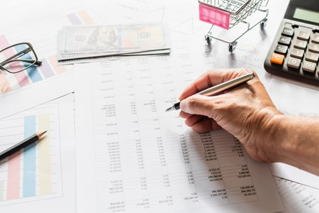 Financial accounting, work at home, Close-up Man\'s hand holding a pen examining accounting numbers. Numbers on paper and calculator, Many numbers, graph, pencil, glasses on paper, Blurred background.の写真素材