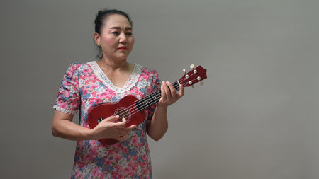 Mature Asian woman enjoys playing small red ukulele. Her floral dress features vibrant pink, blue, white patterns. Looks focused, expressing peaceful musical hobby, leisure time. Studio shot, neutralの写真素材