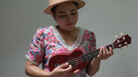 Woman plays red ukulele, focused expression. Straw hat, vibrant floral dress. Music hobby, leisure...
