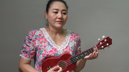 Mature woman playing red ukulele. Floral dress displays pink, blue, white flowers. Person concentrating, enjoying music. Hands strumming chords. Serene portrait. Musical instrument hobby. Leisureの写真素材
