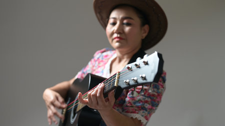Mature woman plays guitar, donning cowboy hat, vibrant floral blouse. Music performance, hobby, leisure activity. Fingers skillfully strumming colorful strings, showcasing musical talent. Creativeの写真素材