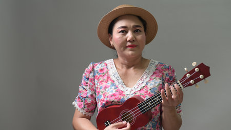 Mature woman, straw hat, floral dress. Plays red ukulele, musical instrument. Hobby, leisure, entertainment. Adult female musician performs. Happy, expressive. Studio background, gray wall. Culturalの写真素材