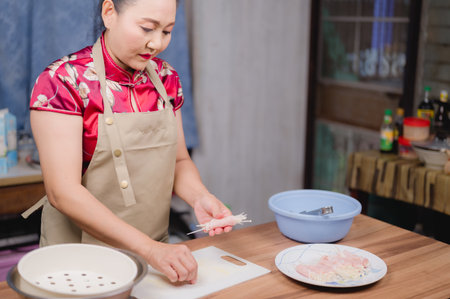 An Asian woman in traditional attire and an apron diligently prepares a healthy meal with fresh ingredients in her bright, cozy home kitchen.の写真素材